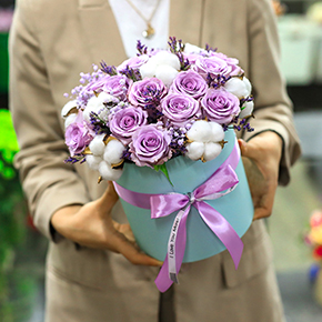 Композиция в шляпной коробке из лиловых роз Меритим, гипсофил и хлопка в Лосино-Петровском